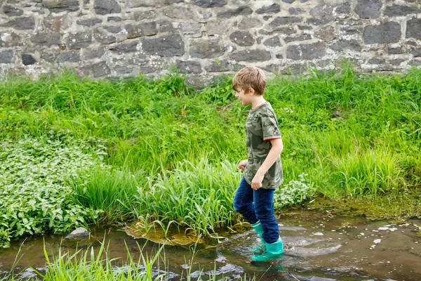 Menino pequeno feliz das crianças da escola que se diverte andando através da água no rio em botas de borracha da gengiva. retrato infantil de criança saudável em férias de verão, jogos ativos ao ar livre — Fotografia de Stock