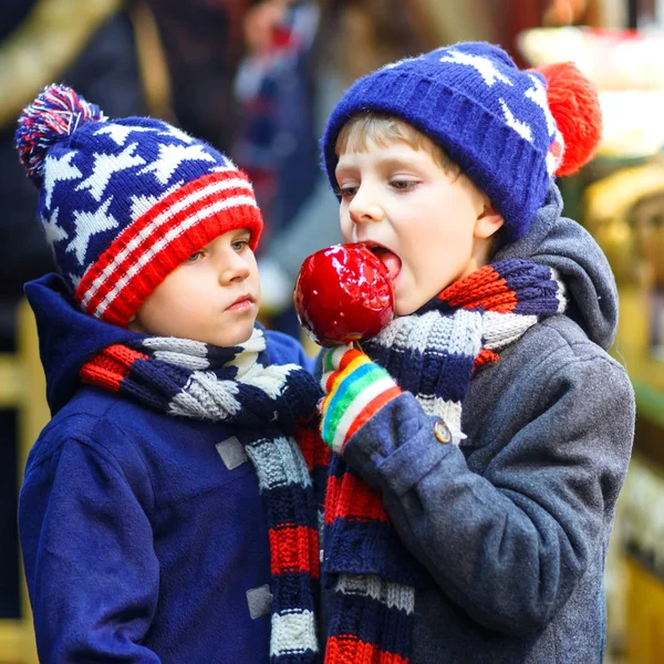 Dos niños pequeños, hermanos lindos comiendo manzana azucarada cerca de un puesto dulce con pan de jengibre y nueces. Niños felices en el mercado de Navidad en Alemania. El ocio tradicional en Navidad —  Fotos de Stock