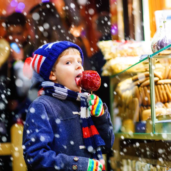 Kleiner süßer Junge isst Zuckerapfel in der Nähe von Süßwarenstand mit Lebkuchen und Nüssen. Glückskind auf dem Weihnachtsmarkt in Deutschland. Traditionelle Freizeit an Weihnachten. Urlaub, Feier, Kindheit — Stockfoto