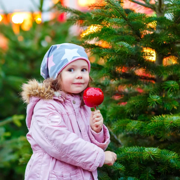 Linda niña comiendo manzana azucarada en el mercado de Navidad. Niño feliz en ropa de moda de invierno con luces en el fondo — Foto de Stock