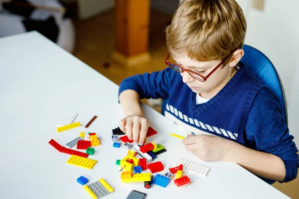 Gözlüklü küçük sarışın çocuk bir sürü renkli plastik blokla oynuyor. Sevimli okul çocuğu robot yaparak ve yaratarak eğleniyor. Yaratıcı eğlence modern teknik ve robotik. — Stok fotoğraf