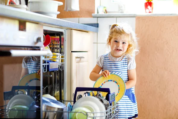 Маленькая милая малышка помогает разгрузить посудомоечную машину. Смешное счастливое дитя, стоящее на кухне, держащее тарелки и кладущее миску на голову. Здоровый ребенок дома. Великолепный помощник веселится — стоковое фото