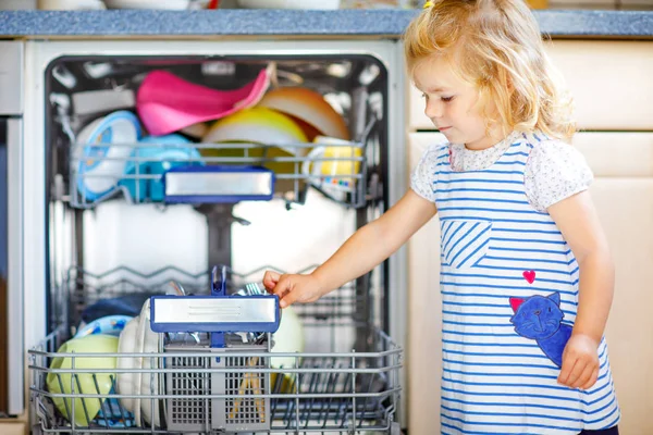 Pequeña adorable linda niña ayudando a descargar lavavajillas. Gracioso niño feliz de pie en la cocina, sosteniendo los platos y poniendo un tazón en la cabeza. Un niño sano en casa. Hermoso ayudante divertirse — Foto de Stock