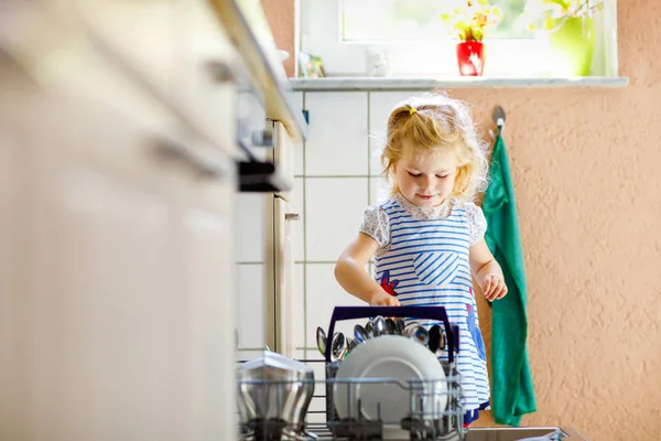 Little adorable cute toddler girl helping to unload dishwasher. Funny happy child standing in the kitchen, holding dishes and putting a bowl on head. Healthy kid at home. Gorgeous helper having fun — Stock Photo, Image