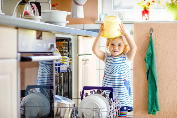 食器洗い機をアンロードするのに役立つ小さな愛らしい幼児の女の子。台所に立つ面白い幸せな子供は、料理を保持し、頭の上にボウルを置く。家にいる健康な子供。楽しさを持つ豪華なヘルパー — ストック写真
