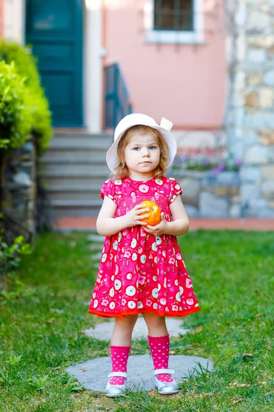 Portretul frumoasei superbe fetițe minunate în roz de vară arată haine, rochie de modă, șosete de genunchi și pălărie. Fericit copil sănătos în fața casei colorate care ține portocaliu în mâini — Fotografie, imagine de stoc