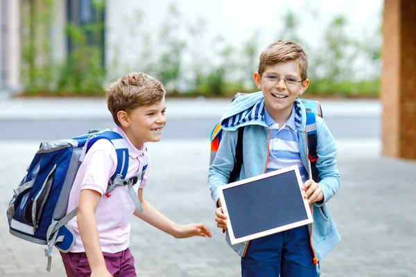 Två små pojkar med ryggsäck eller väska. Skolbarn på väg till skolan. Friska barn, bröder och bästa vänner utomhus på gatan lämnar hemmet. Skolor på kritskrivbordet. Lyckliga syskon. — Stockfoto