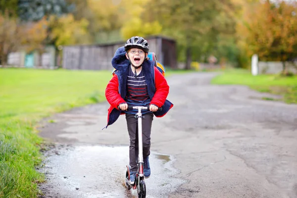 Carino ragazzino della scuola cavalcando su scooter push sulla strada da o per la scuola. Studente di 7 anni che guida attraverso pozzanghere. divertente bambino felice in abiti colorati di moda e con casco. — Foto Stock
