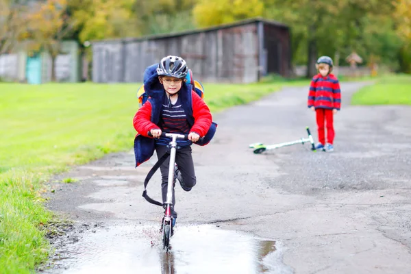 Two little kids boys riding on push scooters on the way to or from school. Schoolboys of 7 years driving through rain puddle. Funny siblings and best friends playing together. Children after school — Stock Photo, Image