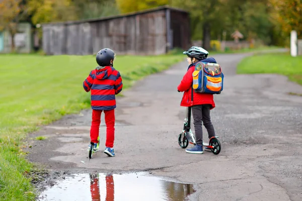 Dos niños pequeños viajando en patinetas de camino a la escuela o desde ella. Colegiales de 7 años conduciendo a través del charco de lluvia. Divertidos hermanos y mejores amigos jugando juntos. Escuela de niños fuera. — Foto de Stock