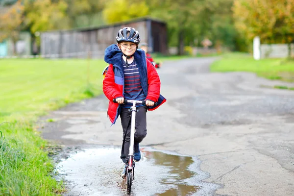 Okula giderken ya da dönerken scooter 'a binen şirin bir okul çocuğu. Yedi yıllık öğrenci yağmur birikintisinin içinden geçiyor. renkli elbiseli ve kasklı komik mutlu çocuk.. — Stok fotoğraf