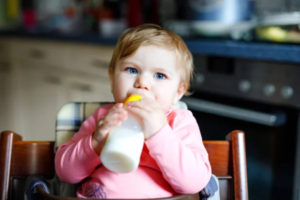 Linda menina adorável segurando mamadeira e bebendo leite fórmula. Primeira comida para bebés. Criança recém-nascida, sentada na cadeira da cozinha doméstica. Bebês saudáveis e conceito de alimentação por mamadeira — Fotografia de Stock