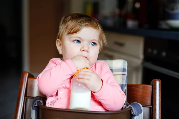 Linda menina adorável segurando mamadeira e bebendo leite fórmula. Primeira comida para bebés. Criança recém-nascida, sentada na cadeira da cozinha doméstica. Bebês saudáveis e conceito de alimentação por mamadeira — Fotografia de Stock