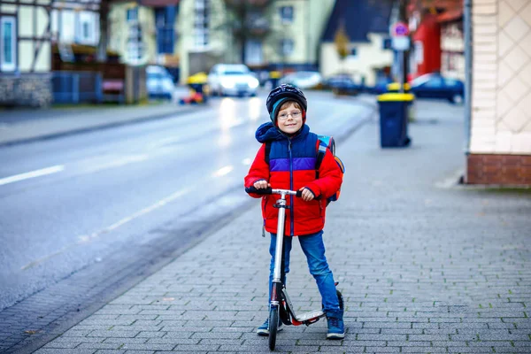 Hermoso niño de escuela montado en scooter camino a la escuela primaria. Niño con casco de seguridad, bolso escolar en otoño lluvioso o mañana fría invierno. Tráfico en la ciudad y escolares —  Fotos de Stock