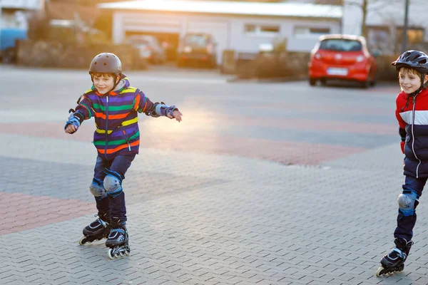 2人の小さな子供たちが市内でローラースケートをしています。幸せな子供たち、兄弟や保護安全服の親友。現役の男子学生がスポーツをしたり、インラインスケートでスケートを学ぶ. — ストック写真
