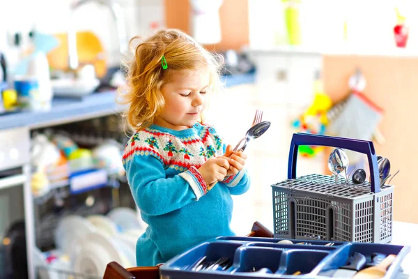 Cute little toddler girl helping in the kitchen with dish washing machine. Happy healthy blonde child sorting knives, forks, spoons, cutlery. Baby having fun with helping housework mother and father. — Stock Photo, Image