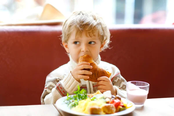 可爱的健康的男孩吃羊角面包和喝草莓奶昔在咖啡馆。快乐的孩子与父母一起吃早餐或在托儿所。蔬菜、鸡蛋作为儿童的健康食品. — 图库照片