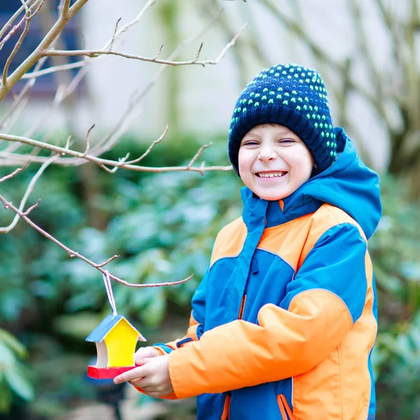 Niño pequeño alimentando pájaros en invierno. Lindo niño preescolar feliz colgando colorido casa de aves hechas a sí mismo en el árbol en el frío día helado. Preescolar en ropa wam colorido. Naturaleza, empatía con los animales . —  Fotos de Stock