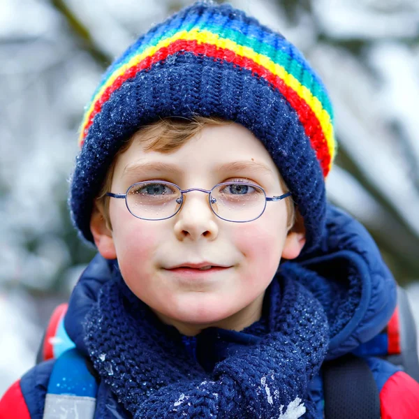 小学生の少年は雪の中学校に歩いている。雪だるまの背景雪と遊んで楽しい子供。子供で目眼鏡バックパックでカラフルな冬服 — ストック写真