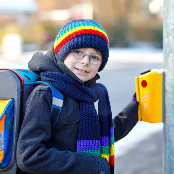 Kleine schooljongen van de lagere klasse die naar school loopt tijdens de sneeuwval. Gelukkig gezond kind met bril drukknop voor verkeerslichten. Met rugzak of tas in kleurrijke winterkleding. — Stockfoto