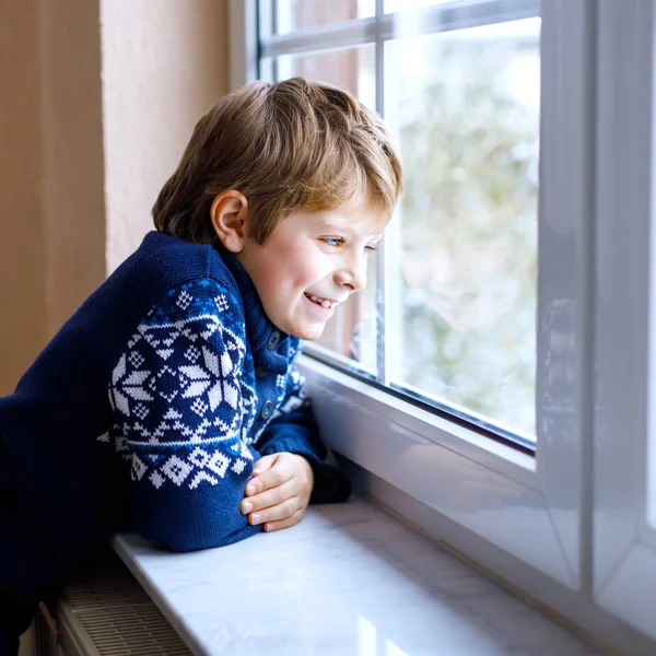 Mutlu sevimli çocuk pencerenin yanında oturuyor ve Noel sabahı ya da sabahleyin dışarıda karları seyrediyor. Gülümseyen çocuk kar yağışı ve büyük kar taneleriyle büyülenmiş..