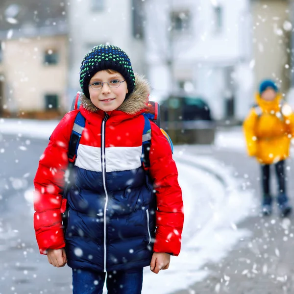 Liten skola unge pojke på elementär klass går till skolan under snöfall. Glada barn och studerande med glasögon ryggsäck i färgglada vinterkläder. schoolkid i gul jacka på bakgrund — Stockfoto