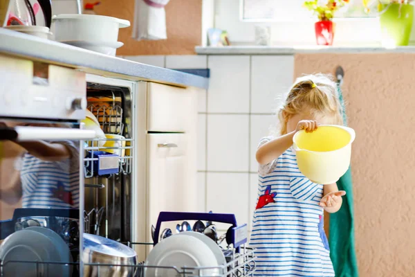 A kis aranyos, aranyos kislány segít kipakolni a mosogatógépet. Vicces, boldog gyerek áll a konyhában, edényeket tart és tálat tesz a fejére. Egészséges gyerek otthon. Gyönyörű segítő szórakozás — Stock Fotó