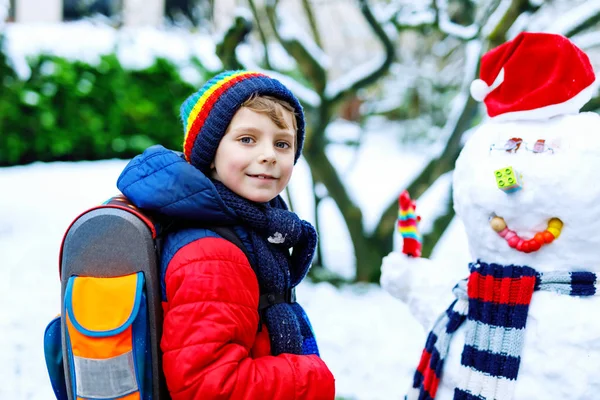 Petit garçon de l'école en vêtements colorés, avec des lunettes et un sac à dos s'amusant avec bonhomme de neige après la fin de l'école primaire — Photo