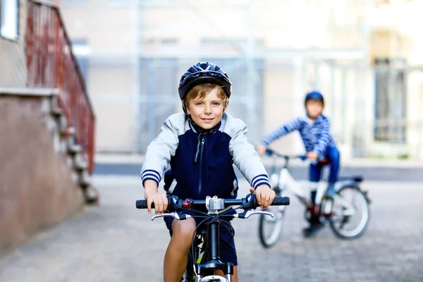 两个戴着安全帽，背着背包骑自行车的学童在城里。穿着五颜六色衣服的快乐儿童骑自行车上学。儿童在户外上学的安全方法 — 图库照片
