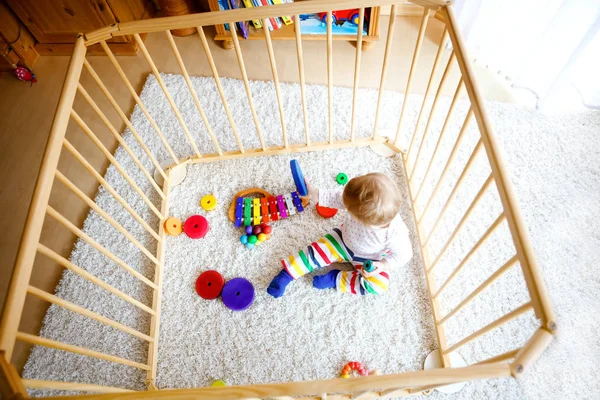 Gyönyörű kislány áll a járókában. Aranyos aranyos gyerek játszik színes játékokkal. Otthon, vagy gyerekszoba, a gyerekek biztonsága. Egyedül a baba vár anyára. — Stock Fotó