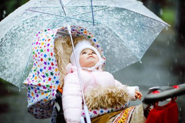 Tatlı küçük güzel kız bebek arabasında oturuyor ya da soğuk bir günde pusetli, yağmurlu ve karlı bir bebek arabasında. Sıcak giysili mutlu bir çocuk, şık bir bebek ceketi. Büyük şemsiyeli bebek