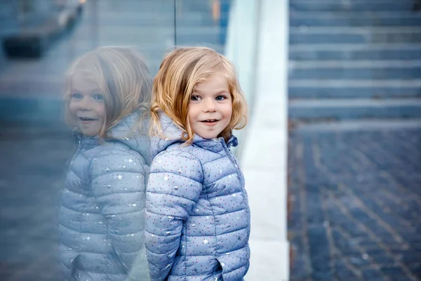 Όμορφο πορτρέτο του χαριτωμένου μικρού κοριτσιού. Χαρούμενο παιδάκι που κοιτάει την κάμερα και χαμογελάει. Με αντανακλάσεις παιδιών στο παράθυρο. Έξω στο σούρουπο. — Φωτογραφία Αρχείου