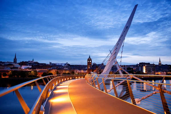 Derry, Ireland. Illuminated Peace bridge in Derry Londonderry, City of Culture, in Northern Ireland with city center at the background. Night cloudy sky with reflection in the river at the dusk — Stock Photo, Image