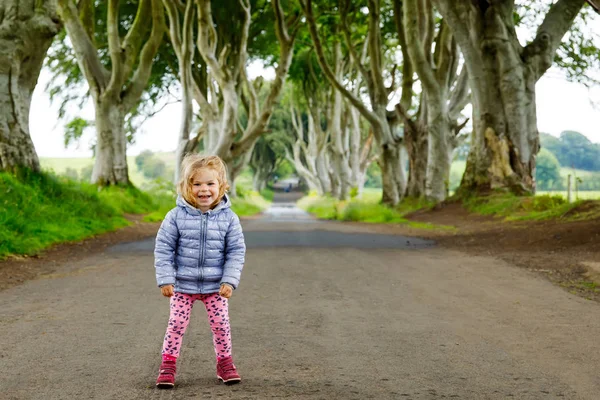 Menina bonito criança andando em um dia chuvoso no início de The Dark Hedges. Irlanda do Norte. Criança feliz visitando com pais e família famosa avenida árvore irlandesa — Fotografia de Stock