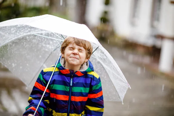 Gyönyörű kisfiú, aki esőben, esőben és hóban sétál az iskolába egy esernyővel a hideg napon. Boldog és örömteli gyermek színes divat alkalmi ruhák. — Stock Fotó