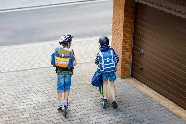 Güneşli bir günde sırt çantası ile şehirde scooter ile sürme güvenlik kask iki okul çocuk çocuklar. Okula giderken renkli kıyafetlerle bisiklete binen mutlu çocuklar. Arkadan tanınmayan yüzler — Stok fotoğraf