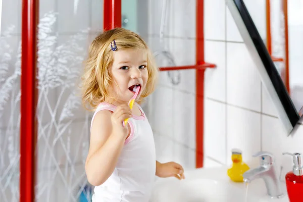 Cute adorable toddler girl holding toothbrush and brushing first teeth in bathroom after sleeping. Gorgeous baby child learning to clean milk tooth. Morning healthy hygiene routine for children