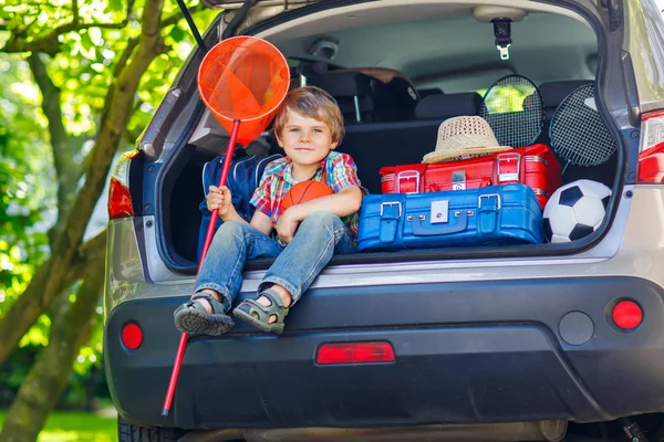 Adorable niño pequeño sentado en el maletero del coche justo antes de salir de vacaciones de verano con sus padres. Niño feliz con maletas y juguetes en viaje — Foto de Stock