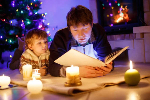 Far och söt liten pojke läser bok med skorsten, ljus och öppen spis. — Stockfoto