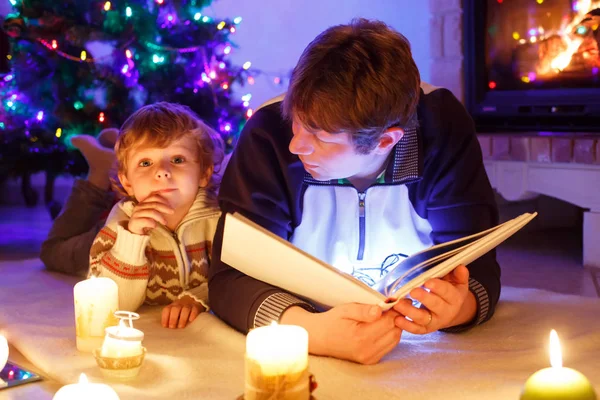 Father and cute little toddler boy reading book by chimney, candles and fireplace. Stock Picture