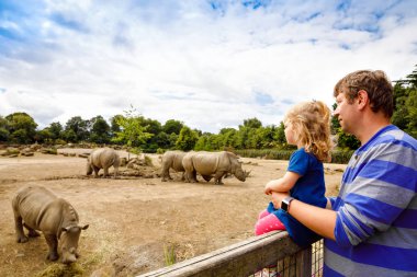 Sevimli sevimli yürümeye başlayan kız ve baba hayvanat bahçesinde gergedanlar izliyor. Mutlu bebek çocuk, kızı ve baba, sıcak yaz gününde hayvanlar safari parkı ile birlikte eğlenen aile.