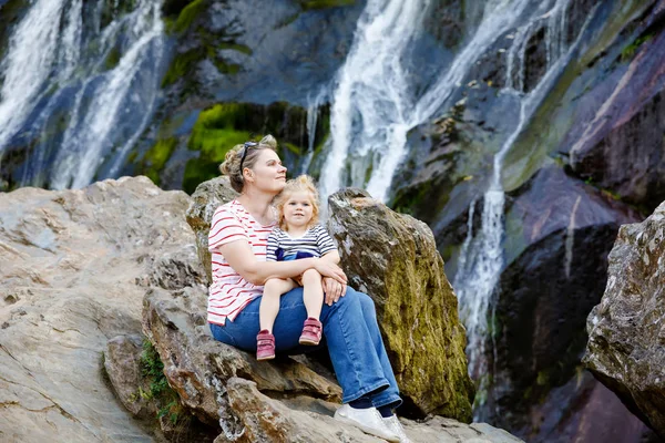 Roztomilá holčička a matka sedí u vodní kaskády Powerscourt vodopádů, největšího vodopádu v Irsku v co. Wicklow. Rodinné dovolené s malými dětmi. Ženská a dětské dítě — Stock fotografie