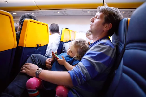 Молодой отец держит свою дочку-малыша во время полета на самолёте, отправляясь в отпуск. Уставший мужчина и симпатичная девушка во время ночного полета. ребенок играет со смартфоном. Семья сидит в самолете — стоковое фото