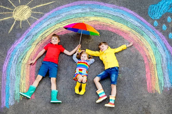 Drei kleine Kinder, zwei Schulkinder Jungen und ein Kleinkind amüsieren sich mit Regenbogenbildern mit bunten Kreiden auf Asphalt. Geschwister in Gummistiefeln malen auf dem Boden und spielen zusammen. — Stockfoto