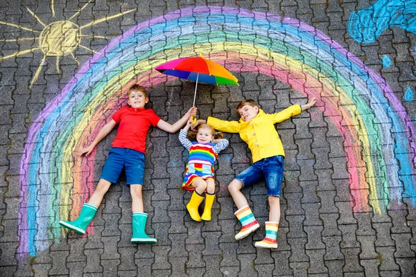 Três crianças pequenas, dois meninos das crianças da escola e menina da criança que se divertem com com desenho da imagem do arco-íris com giz colorido no asfalto. Irmãos em botas de borracha pintando no chão brincando juntos . — Fotografia de Stock