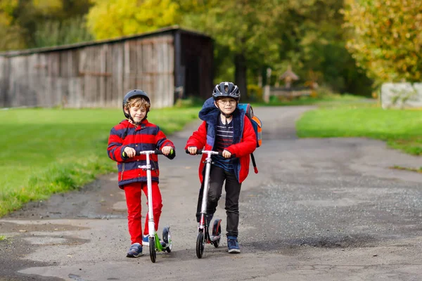 Twee jongetjes rijden op scooters op weg naar of van school. Schooljongens van 7 jaar die door een regenplas rijden. Grappige broers en zussen en beste vrienden die samen spelen. Kinderen naar school. — Stockfoto