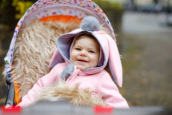 Schattig klein mooi babymeisje zittend in de kinderwagen of kinderwagen op herfstdag. Gelukkig lachend kind in warme kleren, mode stijlvolle roze babyjas met konijnenoren. Baby gaat wandelen met ouders. — Stockfoto