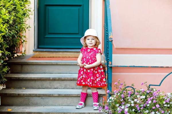 Porträtt av vacker liten gorgeus härlig småbarn flicka i rosa sommar titta kläder, mode klänning, knästrumpor och hatt. Glad frisk baby barn poserar framför färgglada hus. — Stockfoto