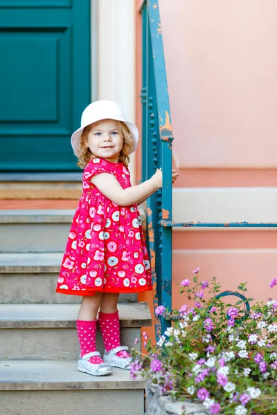 Portret van prachtige kleine gorgeus mooie peuter meisje in roze zomer look kleding, mode jurk, knie sokken en hoed. Gelukkig gezond baby kind poseren voor de voorkant van kleurrijke huis. — Stockfoto