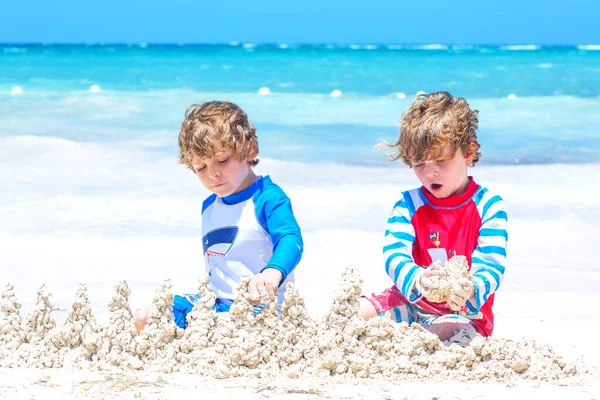 İki küçük çocuk adada tropikal plajda kumdan kale inşa ederken eğleniyor. Tatillerinde birlikte oynayan sağlıklı çocuklar. İkizler, Mutlu kardeşler gülüyor ve gülümsüyor — Stok fotoğraf